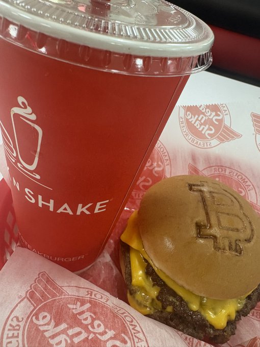 A close-up of a Steak 'n Shake meal featuring a red milkshake cup and a cheeseburger with the 'S' logo branded on the bun, resting on branded paper.