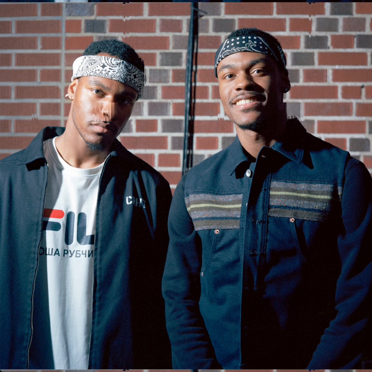 Two young men wearing bandanas and dark jackets pose against a brick wall, with the man on the left wearing a white Fila t-shirt featuring Cyrillic text ("РУБЧИ"). The image relates to the post by showcasing the twin subjects of Justin Aversano and Kim Asendorf's "Doppelgänger #256" NFT, a piece from their project that pairs photographs of unrelated people who share strikingly similar appearances, with this particular edition sold on Verse Secondary for $271.