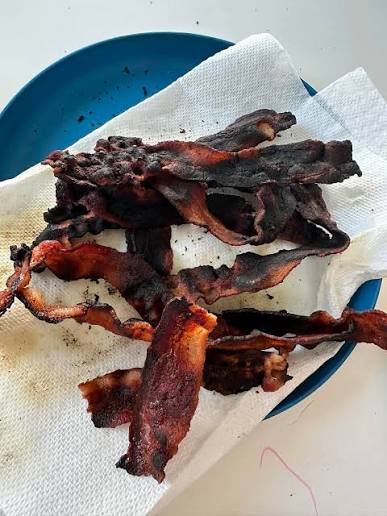 Several pieces of burnt, overcooked bacon rest on a paper towel-lined blue plate on a white surface.