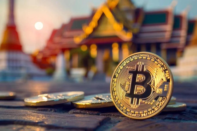 A golden physical Bitcoin token resting on top of a pile of gold coins on a paved surface, with a blurred temple-like building and sunset in the background.
