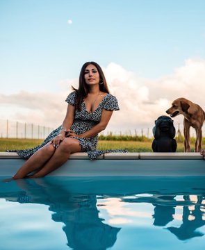 A young woman in a floral dress sits on the edge of a pool, posed with two dogs against a sunset sky with the moon visible, complementing the post's message of "Living inside the moment" shared with the NFT community on objkt.com.