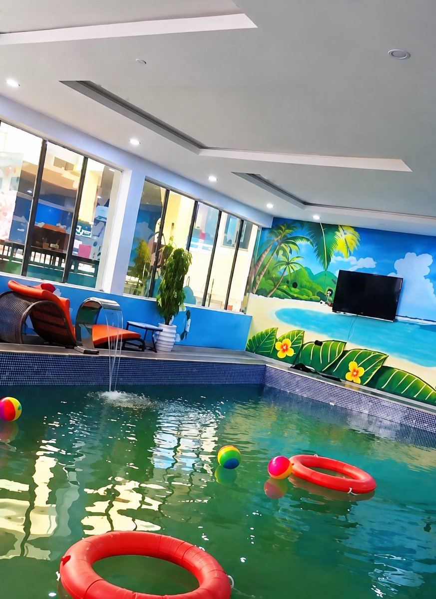 An indoor swimming pool with green-tinted water featuring red life rings, colorful beach balls, a waterfall feature, lounge chairs, and a tropical beach mural on the wall with a mounted TV.