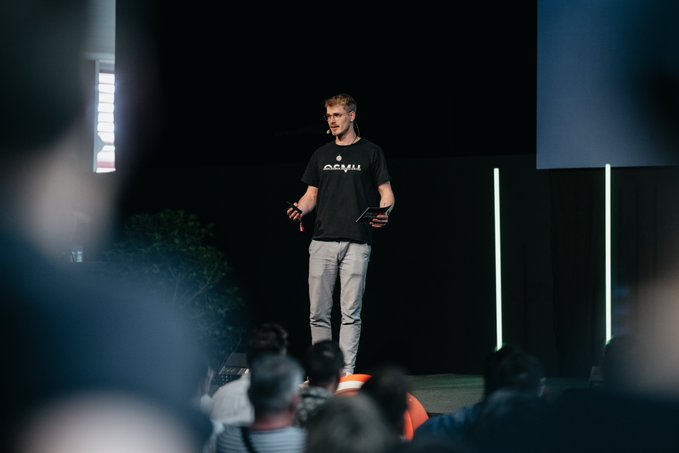 The image shows a man standing on stage, giving a speech or presentation to an audience. The speaker appears to be wearing a black shirt with white text that reads "Last year @BTCPrague was just amazing! Make sure to give it a go this year". The background of the image is dark and out of focus, drawing attention to the man speaking on stage.