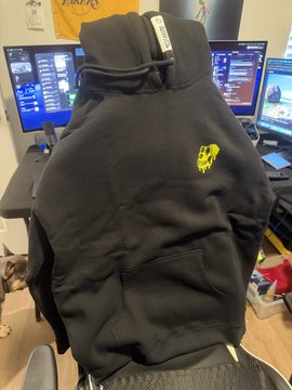 A black hoodie with a yellow logo is draped over a chair in front of a dual-monitor gaming/computer setup.