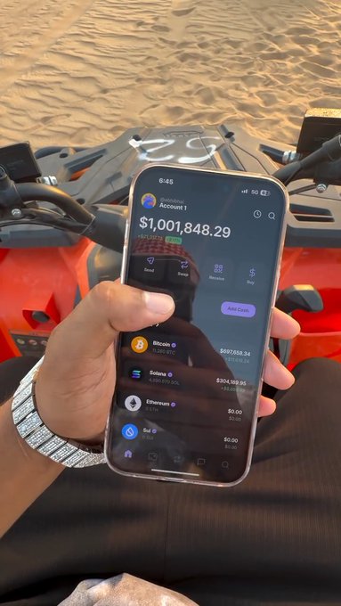 A person holding a smartphone showing a crypto portfolio worth $1,001,848.29 in a Coinbase account, with Bitcoin and Solana positions visible, while sitting on what appears to be an ATV in a desert setting.