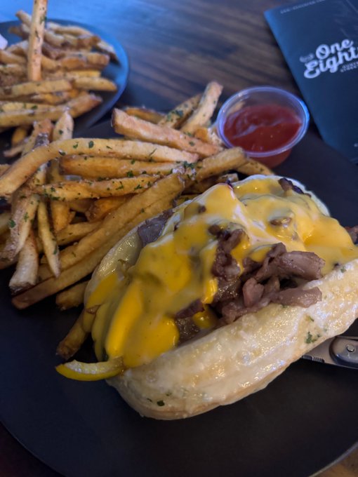A Philly cheesesteak loaded with melted cheddar, beef, peppers, and onions sits alongside a generous plate of seasoned fries and ketchup. In the background, a menu reads "One Eight," suggesting a stop at the One Eight restaurant for a casual meal.
