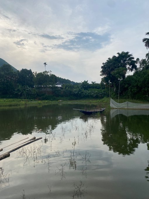 A serene landscape photograph of a calm pond reflecting the sky, with a small wooden boat floating near the shore, lush green trees, and mountains in the background.