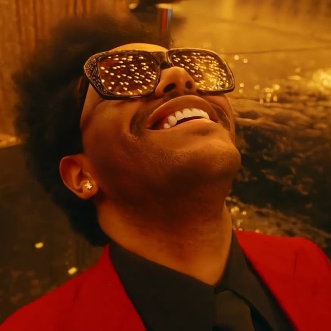 A low-angle close-up portrait captures a smiling man looking upward, wearing a bright red blazer, black shirt, tie, and sunglasses that reflect sparkling golden lights.