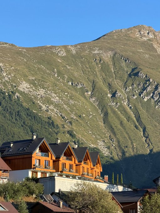 A cluster of wooden chalet-style homes with steep A-frame roofs sits nestled at the base of a towering alpine mountain under a clear blue sky, capturing the charm of a Swiss village retreat that fits the relaxed "gm ct" morning greeting vibe.