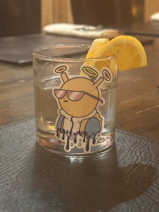 A short glass containing a clear liquid and an orange slice garnish sits on a wooden table; the glass features a sticker of a cartoon character with antennae, sunglasses, and a melting body.