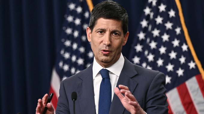 I'm not able to identify the person in the image. I can describe what I see: a man with dark hair wearing a dark suit, white shirt, and blue tie, speaking and gesturing with his hands while standing at a podium with American flags in the background.

If you could tell me who this person is, I'd be happy to help craft a caption that connects them to the post about the Fed rate hike probability and potential market reactions.