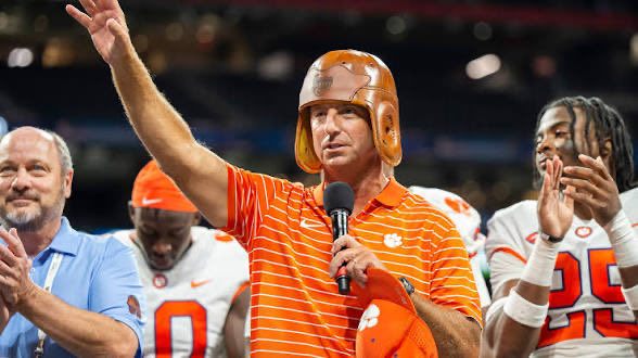 A photograph of a man in an orange and white striped shirt and helmet speaking into a microphone with his arm raised, standing with a football team on a field.