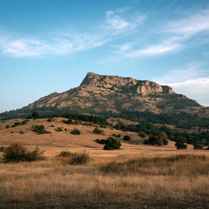 Scenic landscape photograph of a rocky mesa or butte rising above golden grasslands dotted with green shrubs, under a bright blue sky with wispy clouds.