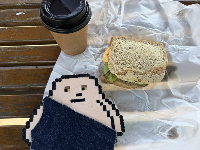 A paper cutout figure with pixel-art styling holds a sandwich and sits beside a takeaway coffee cup on white paper atop a wooden slatted surface.