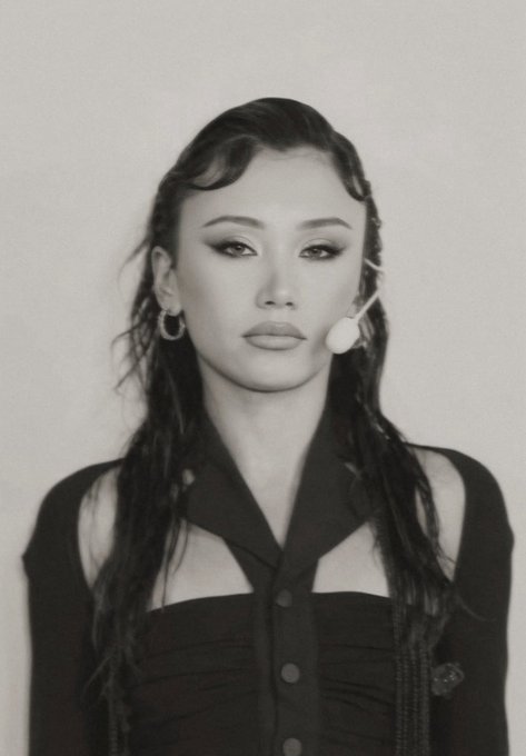 A black and white portrait photograph of a young woman with dark hair, wearing a black cut-out top and hoop earrings, wearing a small headset microphone.