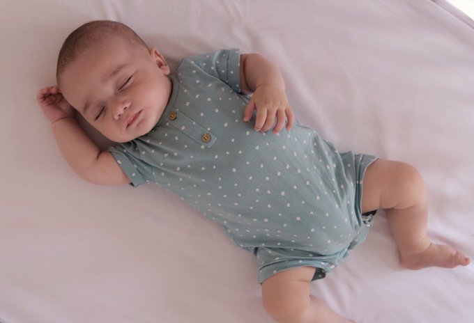 A sleeping baby wearing a sage green polka-dot onesie lies on a white sheet.