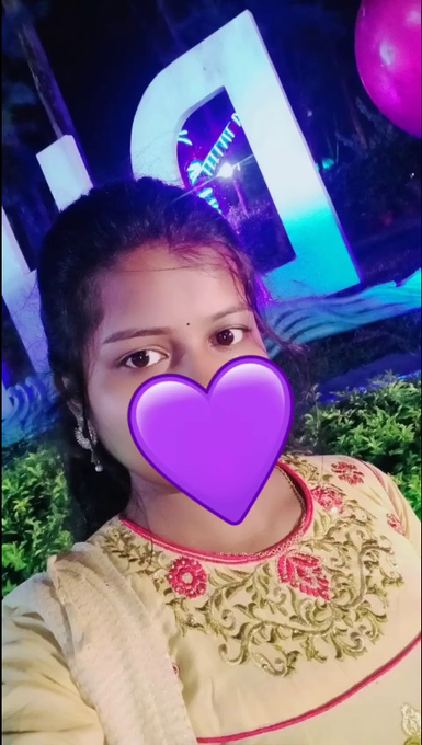 Selfie of a young woman in traditional embroidered Indian attire with a purple heart emoji covering her mouth, taken in front of illuminated signage at night.