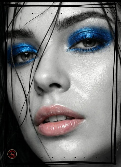 A high-contrast black-and-white close-up portrait of a woman with dramatic blue eyeshadow, wet hair strands across her face, and glossy pink lips, framed by a thin black border with small dots.
