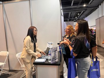 The image shows three women standing around a table at an event or convention, likely attending a panel discussion or networking session. One woman appears to be presenting something while her companions listen attentively. On the table are two laptops placed next to each other, possibly for use during the presentation or discussion. The background reveals a large room filled with more people and additional tables where similar activities might take place. The image also includes text that references Philippine Blockchain Week, which suggests that this event could be related to the theme of blockchain technology.