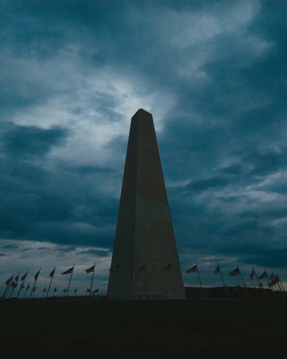 The image shows a tall obelisk, possibly an architectural monument or a memorial, standing prominently against a dark cloudy sky. There are numerous flags hanging around the base of the obelisk that seem to be waving in the wind on this cloudy day. The image also contains several text elements such as the word "UNTIL" and "HISTORY", which might suggest the significance of the monument or memorial.