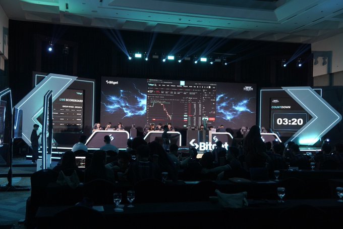 A photograph of a crypto or gaming tournament in a large auditorium, featuring a main screen with a 'Live Scoreboard' and a countdown timer at 03:20.