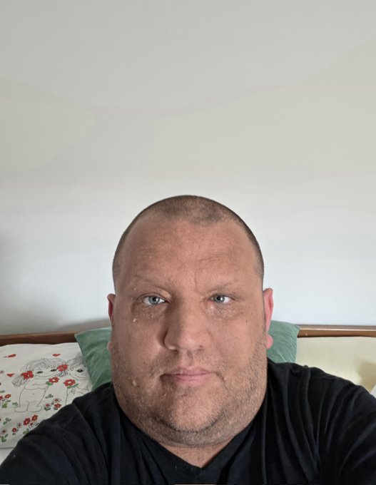 The image features a man with a bald head wearing a black shirt, sitting on a bed against a white wall. The photo has been edited to include a caption that reads "This is the newest pic of your $INJ Boy Dressed in black, like a true ninja Expect the unexpected, very soon P.S. Didn'qt shaved, otherwise I'd look like a kid". This caption adds an element of humor and surprise to the image, making it more engaging for viewers.