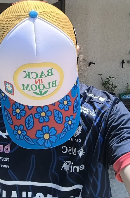 A person wears a vibrant "Bloom in Back" trucker hat with a floral brim and a yellow mesh top, paired with a patterned jersey. The "Gm! Apechain on top!" caption reflects a cheerful morning greeting within the Apechain crypto community, with the colorful, playful hat capturing the fun, casual vibe of the message.