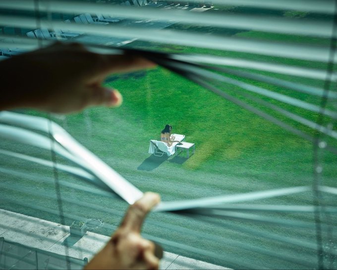 A hand parts venetian blinds to reveal a lone figure in a suit sitting at a desk isolated on an empty green field—a trader peering out at a vast, exposed landscape of risk, much like an investor anxiously watching small market volatility from the safety of their window, unaware that the real danger is the stress itself.