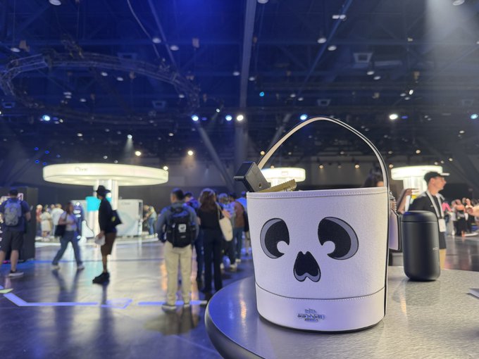 A white Karl Lagerfeld bucket bag with a skull design sits on a table at what appears to be a convention center or exhibition hall, with people mingling in the background under blue lighting.
