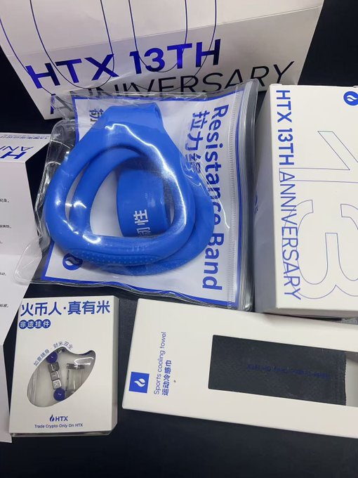 A collection of HTX (Huobi) 13th anniversary merchandise is displayed, including a blue "Resistance Band," branded boxes, and a "Sports cooling towel," alongside Chinese text reading "火币人·真有米" (Huobi people really have rice) and "只有经历." The image visually showcases the promotional swag referenced in the post, highlighting the commemorative items celebrating Huobi's 13-year milestone.