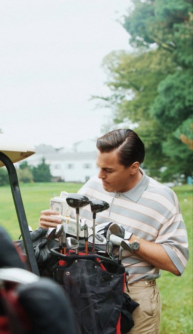 A man dressed in preppy golf attire examines a dollar bill near his golf bag filled with clubs. The image's leisurely, affluent setting ironically contrasts with the post's "gm to those who didn't get liq'd" — a greeting to crypto traders who survived the liquidation wave overnight.