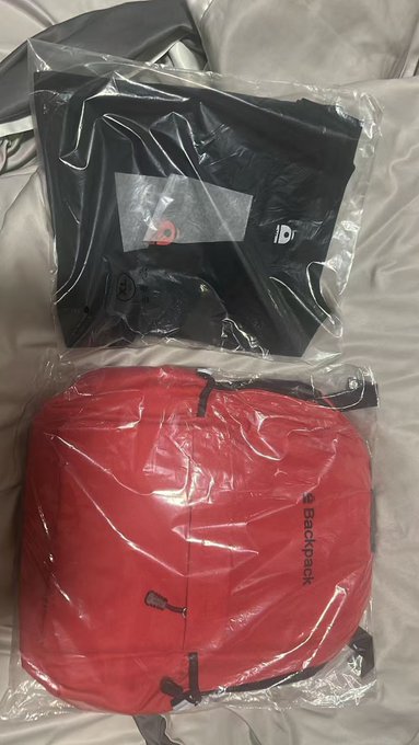 Two items sealed in clear plastic packaging: a black folded garment with a logo, and a red backpack labeled 'Blackpink'.