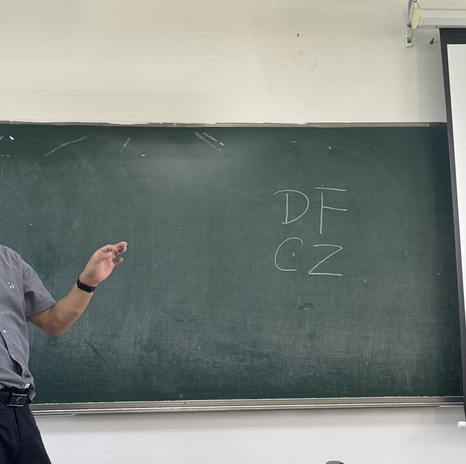 A teacher stands in front of a green chalkboard with the letters "DF" and "CZ" written in white chalk, gesturing with his hand while holding chalk.
