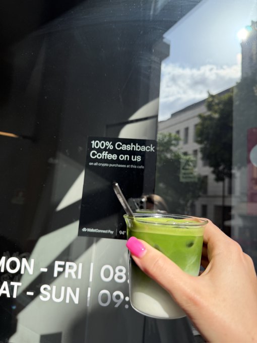 A hand with pink nails holds a layered green and white iced matcha drink with a glass straw outside a cafe featuring a '100% Cashback Coffee on us' WalletConnect Pay sign on the window.