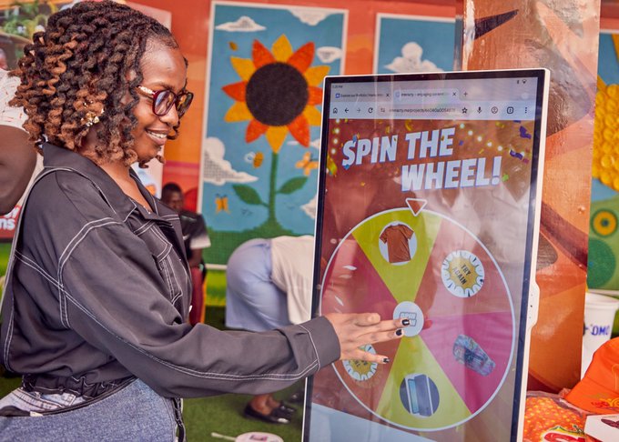 A smiling woman with glasses interacts with a 'Spin the Wheel!' digital kiosk, touching the screen to spin for prizes in a colorful indoor setting.