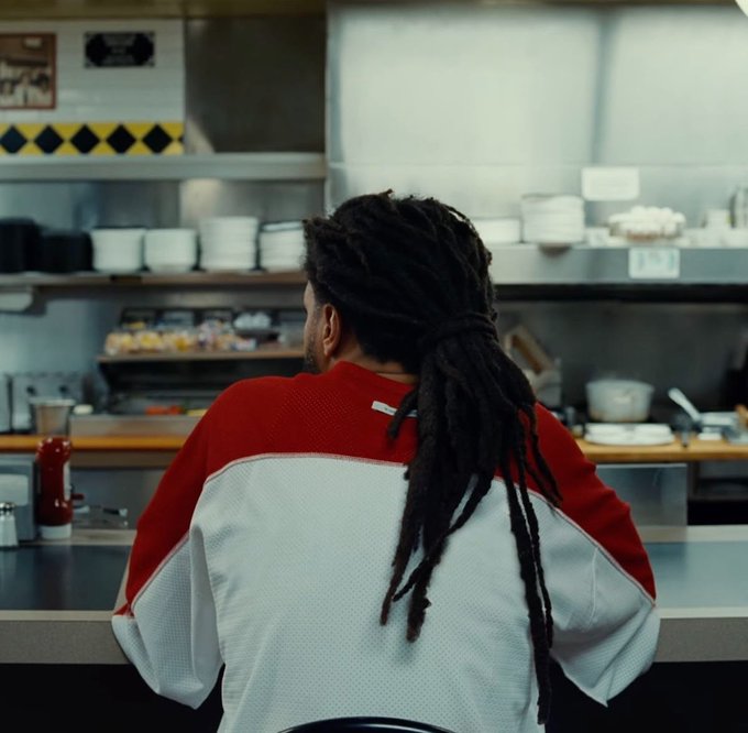 A man with long dreadlocks tied back sits at a diner counter viewed from behind, wearing a red and white color-blocked shirt, facing a commercial kitchen with stainless steel equipment.