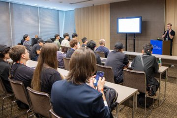 A business presentation or seminar in a conference room with an audience seated facing a presenter standing next to a screen, while attendees take notes or use phones.