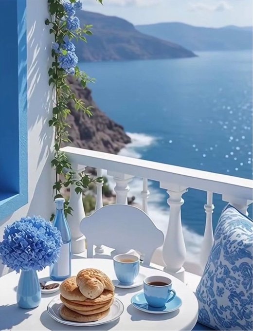 A scenic table setting features blue and white coffee cups, bread, and pastries, accompanied by blue flowers on a balcony overlooking a vast blue ocean and cliffside.