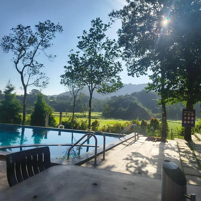 A scenic view from a resort or villa features a swimming pool in the foreground looking out over lush green rice fields and distant mountains under a sunny sky.