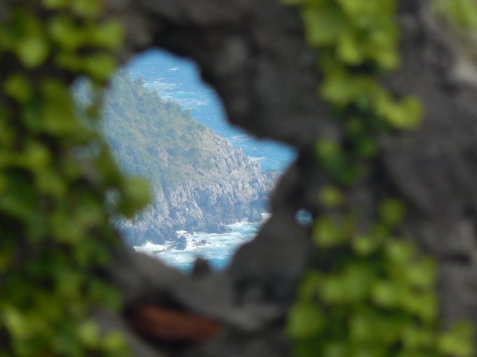 An ocean view of waves crashing against rocky cliffs, seen through a hole framed by out-of-focus dark rocks and green foliage in the foreground.