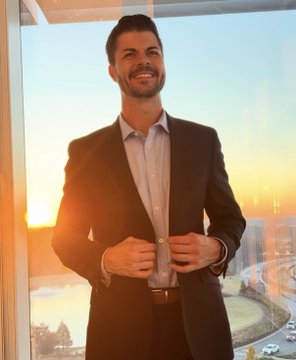 A man with short dark hair and a beard smiles while buttoning his dark suit jacket, standing by a window with a sunset cityscape view behind him.