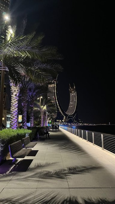 A nighttime view of a waterfront promenade lined with palm trees, with an illuminated twisted skyscraper in the distance.