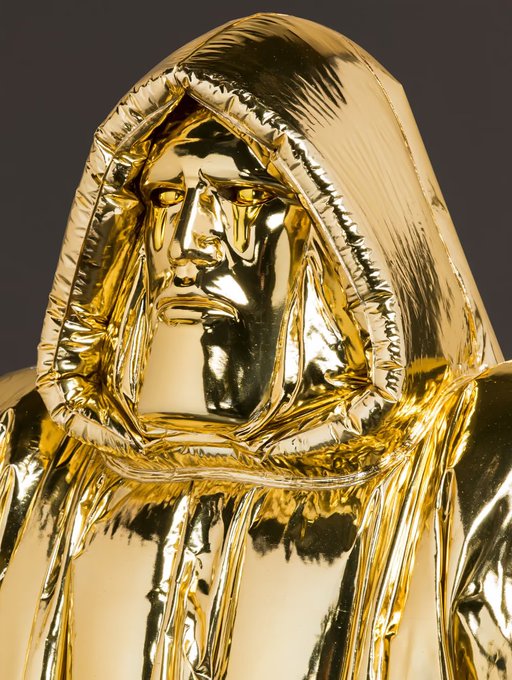 A close-up photograph of a glossy gold-colored sculpture depicting a hooded figure with a downcast, sorrowful facial expression.