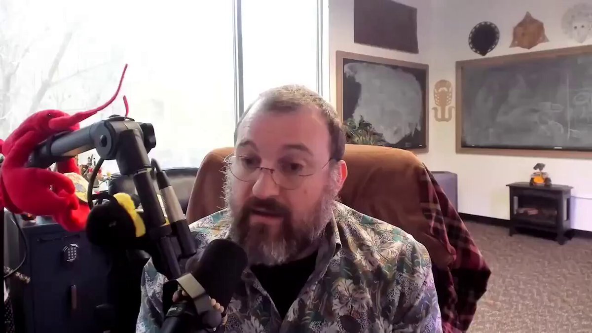 The image shows a man wearing glasses sitting in front of a microphone, possibly participating in an interview or podcast recording session. He has a beard and is dressed in a floral shirt. There are two red stuffed animals placed next to him on his lap, adding a playful touch to the scene. The background features a window with blinds that allow natural light into the room, creating a comfortable atmosphere for the man's conversation.