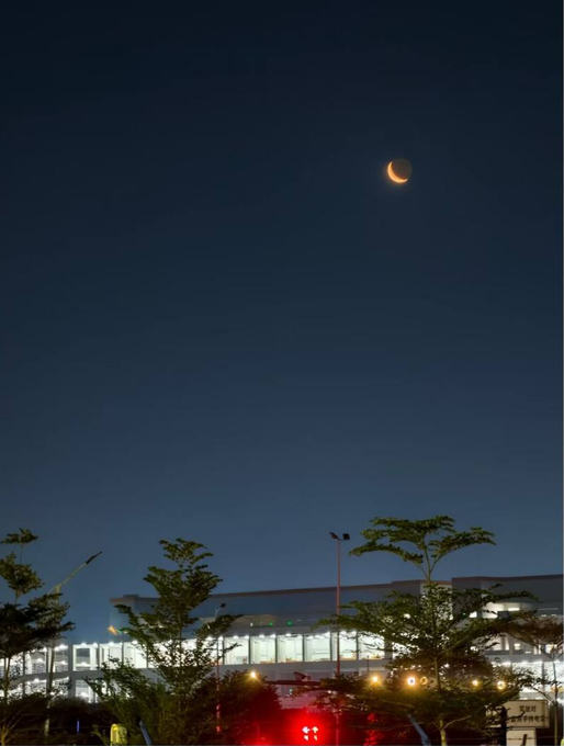 A crescent moon hangs in the dark sky above a brightly lit building and treetops, with no visible text or numbers in the image itself.