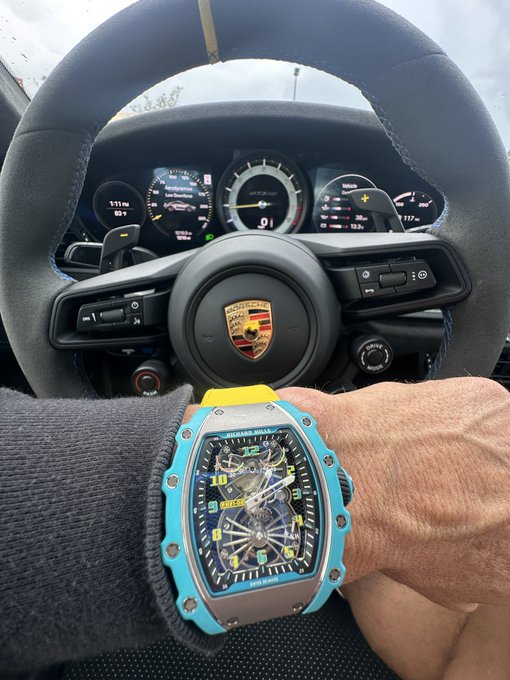 A wrist wearing a vibrant blue Richard Mille watch rests on the steering wheel of a Porsche, with the dashboard displaying 111 mi of range and the central tachometer reading "01." The post's reference to "Hodl" and supply-driven scarcity cleverly ties the exclusive, hard-to-find nature of the luxury timepiece—often only seen on the wrists of the ultra-wealthy—to crypto investment principles.