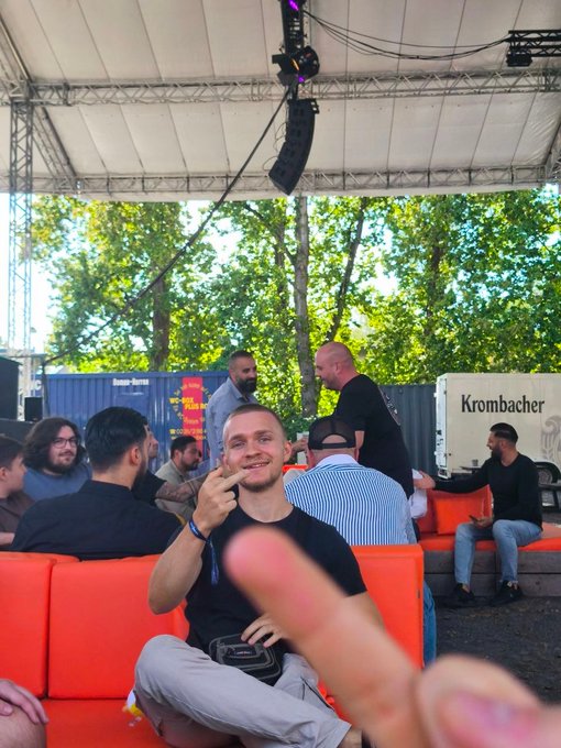 A young man sitting on an orange couch smiles and makes a peace sign at an outdoor event, with a Krombacher truck and other attendees in the background.
