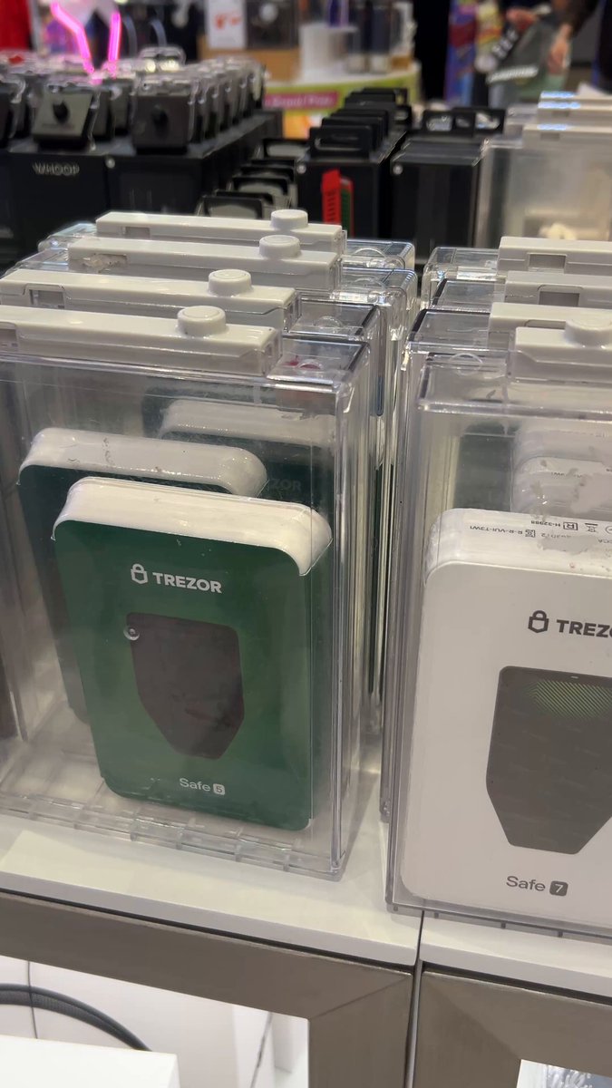 The image shows two clear plastic boxes containing Trezor products sitting side by side on a table. The boxes are filled with various electronic devices such as smartphones, smartwatches, and other accessories. There are also several people present in the background, possibly browsing or shopping for these items.