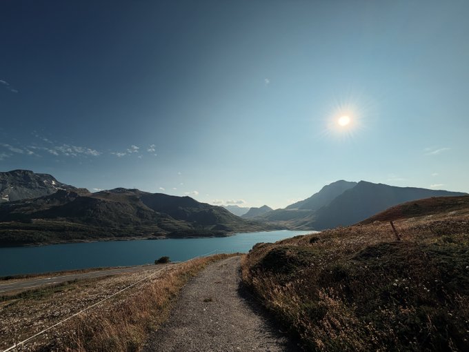 A scenic mountain landscape with a winding dirt path leading down to a serene blue lake, bathed in bright morning sunlight — a fitting visual for a "Gm." greeting, capturing the calm and fresh start of a new day.