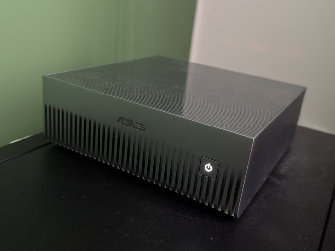 A small form factor black ASUS mini PC sitting on a dark surface against a green and white wall.